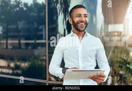Ritratto di un giovane fiducioso sorridente uomo indiano tenendo un tablet e guardando in lontananza Foto Stock