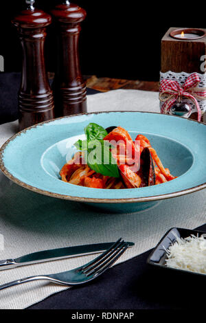 Cucina Italiana. Pasta con pesce e frutti di mare, i pomodori e il formaggio in un blu, una forchetta e un coltello su uno sfondo luminoso. Foto Stock