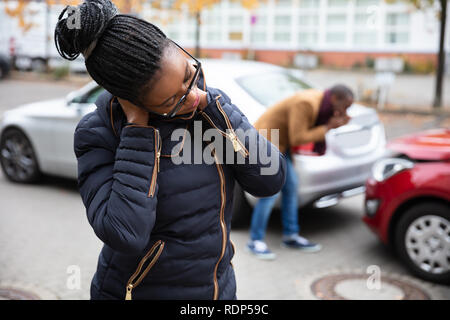 La donna che soffre di dolori al collo di fronte un uomo guarda auto danneggiata su strada Foto Stock