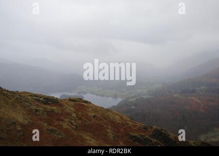 Vista dalla montagna (Loughrigg cadde) giù sul lago di Grasmere in una nebbiosa valle, con le altre montagne in fuga una primavera di nebbia. Colore di autunno Foto Stock
