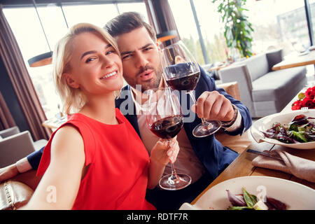 Una giovane coppia in data nel ristorante seduto con i bicchieri di vino rosso cheers tenendo selfie foto gioiosa Foto Stock