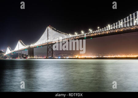 La baia illuminata ponte di collegamento tra San Francisco e Oakland attraverso l'isola del tesoro su una chiara notte' lunga esposizione; San Francisco Bay Area, Californi Foto Stock