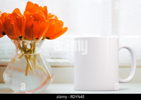 Tazza vuota accanto a un mucchio di tulipani arancio in un vaso di vetro su un davanzale. Perfetto per le imprese che vendono tazze, appena sovrapporre il tuo preventivo o di design o Foto Stock