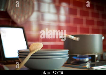 Pila di piatti blu con miscelazione di legno cucchiaio, pentola per la cottura sul fornello a gas e tablet con ricetta. Cucina rosso in background. Foto Stock