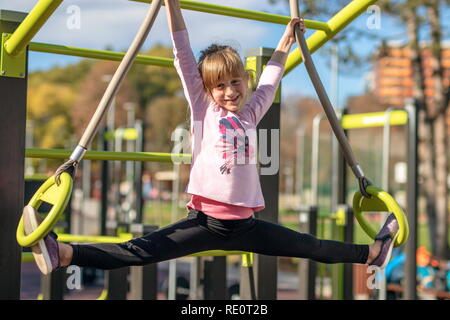 Ragazza giovane si allunga in gruppi in posizione gli anelli di ginnastica al di fuori su di un parco giochi per bambini Foto Stock