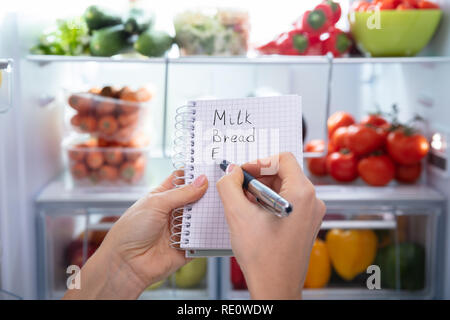 Mano rendendo elenco di cibo sul blocco note a spirale nella parte anteriore di un frigorifero aperto Foto Stock