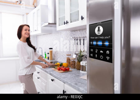 Un forno con la funzione di riconoscimento vocale vicino sorridente giovane donna tagliare le verdure in cucina Foto Stock