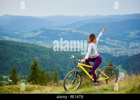 Ragazza sportivo biker ciclismo su giallo in mountain bike con la mano fino a sera Nuvoloso. Montagne, foreste e piccola città sul fondo sfocato. Outdoor sport attività, concetto di stile di vita Foto Stock