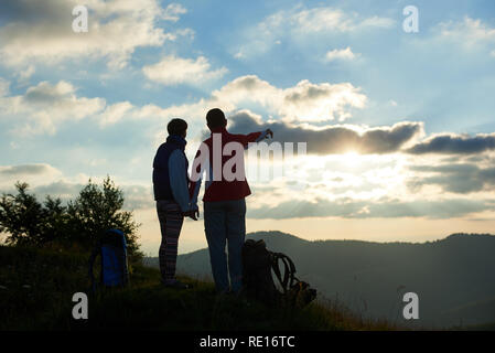 Silhouette di due turisti sono in piedi sulla cima della montagna tenendo le mani vicino zaini contro lo sfondo delle montagne e il cielo nuvoloso al tramonto. Ragazzo che mostra il suo lato della distanza. Vista posteriore Foto Stock