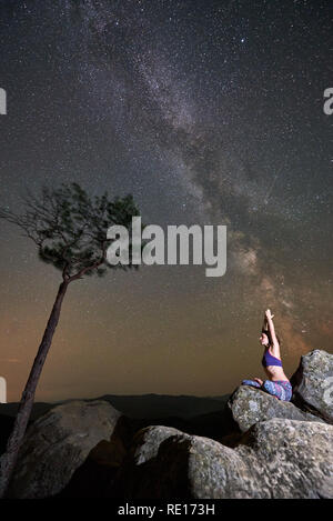 Bella notte estiva in montagna. Donna seduta da sola sulla sommità di un enorme masso fare yoga vicino a pino, sotto il cielo limpido con stelle lampeggianti e la Via Lattea. Turismo ed escursionismo concetto. Siddhasana Foto Stock