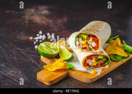 Burrito alla griglia si avvolge con pollo, fagioli, mais, pomodori e avocado su una tavola di legno, sfondo scuro. Carni burrito, cibo messicano. Foto Stock