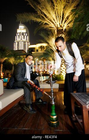 Uomo di fumare una pipa ad acqua shisha,, presso la piscina di sera, il Palace Hotel, Oldtown Dubai, Emirati Arabi Uniti, Medio Oriente Foto Stock