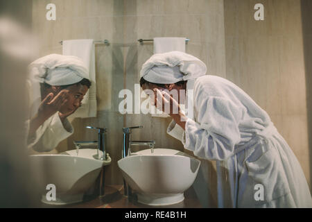 Vista laterale della donna africana in accappatoio lavando la faccia nel lavandino del bagno e guardando nello specchio. Femmina matura la pulizia viso in bagno. Foto Stock