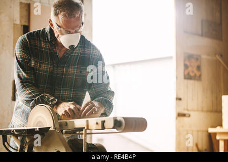 Senior falegname con maschera utilizzando levigatrice a nastro. Carpenter la levigazione di un legno con levigatrice a nastro in falegnameria officina. Foto Stock