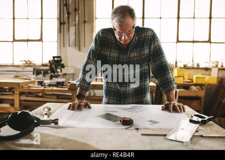 Senior falegname disegno di controllo in officina. Uomo maturo leggendo e studiando la costruzione di piano piano prima di iniziare il suo lavoro in laboratorio di falegnameria. Foto Stock