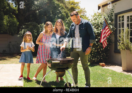 Famiglia avente fun la cottura di cibo insieme nel loro cortile. Uomo che fa un barbeque in piedi nel loro cortile con moglie e figli. Foto Stock