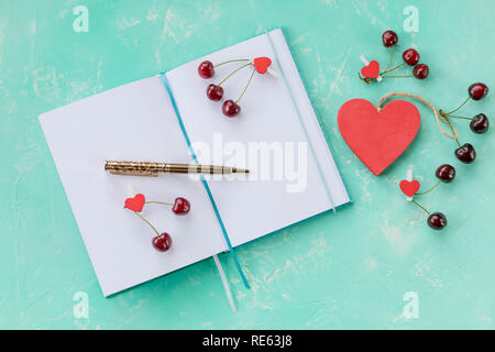 Composizione con il diario e le ciliege dolci e penna, aprire il libro bianco note book e con decor di rosso ciliegia, cuore di legno verde sul tavolo in legno.amore diario e cuori rossi Foto Stock