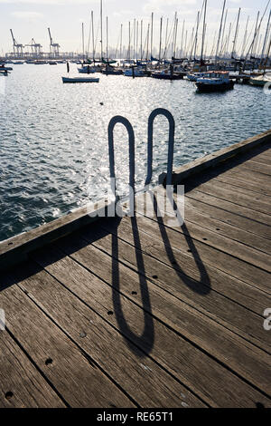 Ombre su un molo in legno dalla parte superiore di una scala che conduce in acqua in una imbarcazione a vela porto. Foto Stock