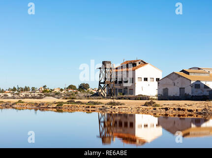 Vecchio edificio rovinato con legno serbatoio acqua sul bordo di un lago salino, entrambi sono riflessi nell'acqua sullo sfondo azzurro del cielo Foto Stock