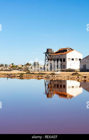 Vecchio edificio rovinato con legno serbatoio acqua sul bordo di un lago salino, entrambi sono riflessi nell'acqua sullo sfondo azzurro del cielo Foto Stock