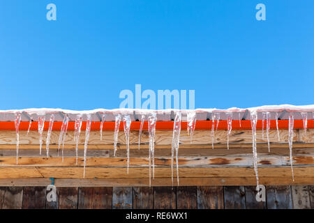 Ghiaccioli appesi ad un tetto durante un inverno canadese. Blue sky contro la costruzione in legno con vernice arancione: comlimentary colori in stile minimalista, con Foto Stock