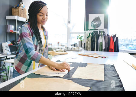 Giovane femmina sarto lavora con schizzi e prodotti tessili e la cucitura di nuovo vestito in design studio Foto Stock