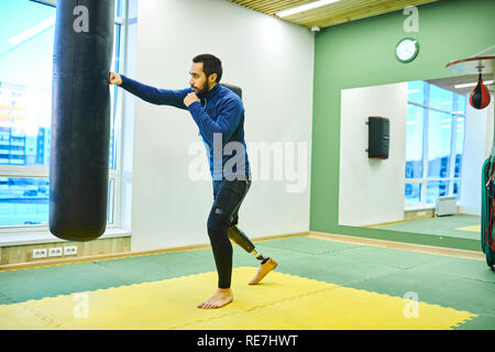 Grave concentrata bello uomo barbuto con gamba protesico di punzonatura Sacco boxe mentre la formazione da soli in palestra con specchio Foto Stock