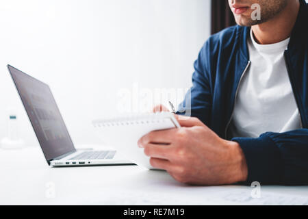 Concentrate l uomo la scrittura di nuove idee in notepad guardando sullo schermo portatile. Giovane collega che siede a tavola e lavoro in ufficio Foto Stock