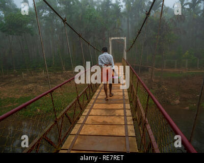 Chikmagalur, India- Dicembre 21, 2018 : sud indiane uomo a camminare in un tradizionale Lungi su un ponte sospeso circondato da noci di betel gli alberi su una nebbia Foto Stock