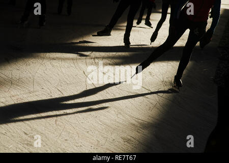 La folla di persone giovani e vecchi insieme divertimento pattinaggio su ghiaccio in inverno al sole con lunghe ombre, motion e sagome Foto Stock