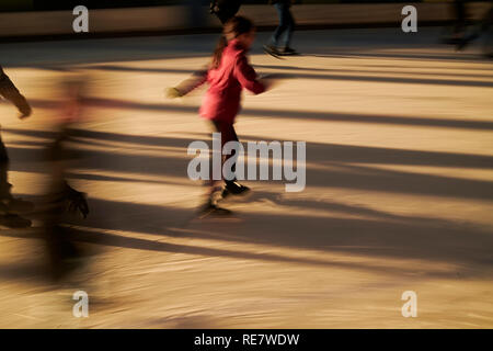 La folla di persone giovani e vecchi insieme divertimento pattinaggio su ghiaccio in inverno al sole con lunghe ombre, motion e sagome Foto Stock