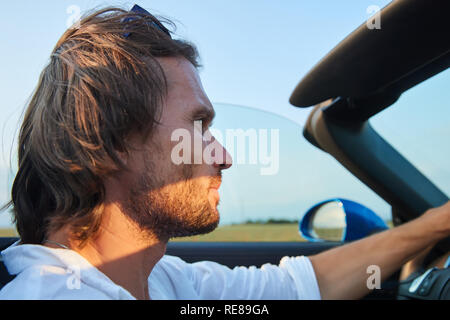 Il giovane uomo brutale con i capelli lunghi e un facile barba di una vettura senza top, cabriolet, colore blu, occhiali da sole Foto Stock