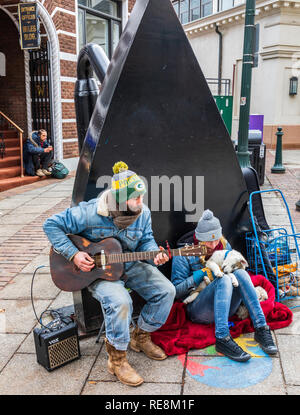 ASHEVILLE, NC, Stati Uniti d'America-1/18/19: un modo di suonare la chitarra busker, accompagnato da una ragazza e due cani con un singolo uomo con zaino seduti sui gradini a sinistra. Foto Stock