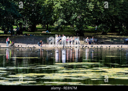 London, Regno Unito - 21 Giugno 2018: St James Park verdi alberi nella soleggiata estate con molte persone camminando sul marciapiede da stagno di acqua di fiume e alimentazione di riflessione Foto Stock