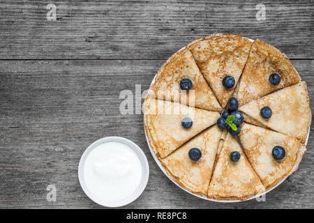 Crepes fatte in casa o sottile frittelle servite con mirtilli freschi e panna acida e menta su tavola in legno rustico. vista superiore con spazio di copia Foto Stock