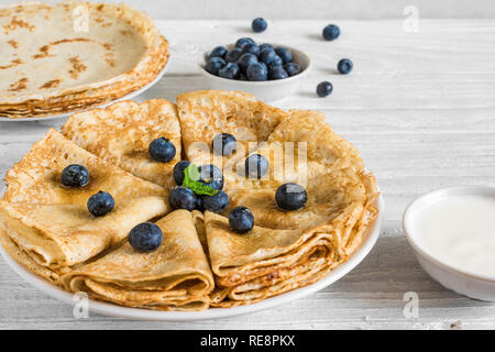 Crepes fatte in casa o sottile frittelle servite con mirtilli freschi, panna acida e menta su bianco tavolo in legno. close up Foto Stock