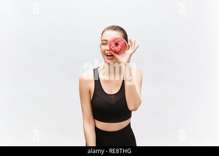 Giovane bella donna sportivo in una canottiera nera pose e sorrisi isolati su sfondo bianco. Ragazza pone con ciambella in mani. Ho ciambella applicato all'occhio Foto Stock