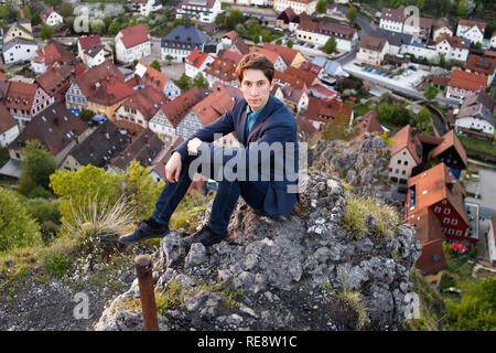 Un uomo in una tuta siede sul ponte di osservazione e guarda la vista della città vecchia di case da sopra il centro storico di Pottenstein in Germania. Foto Stock