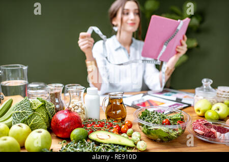 Donna dietologo in uniforme medica lavorando su un piano di dieta seduta con diversi cibi sani ingredienti nel verde office Foto Stock
