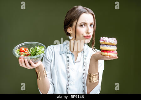 Giovane donna nutrizionista cerca su ciambelle con tristi emozioni scegliendo tra insalata e dessert malsana sullo sfondo verde Foto Stock