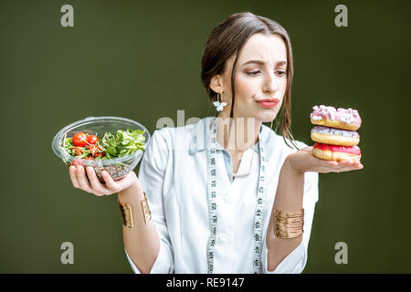 Giovane donna nutrizionista cerca su ciambelle con tristi emozioni scegliendo tra insalata e dessert malsana sullo sfondo verde Foto Stock