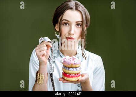 Ritratto di una donna triste nutrizionista in camice con ciambelle e nastro di misurazione sullo sfondo verde. Mangiare malsano e concetto di adiposità Foto Stock