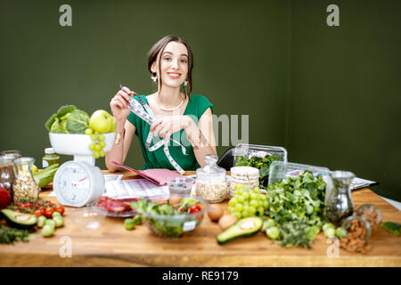 Ritratto di una donna bellissima con nastro di misurazione seduta con vari cibi sani ingredienti sullo sfondo verde Foto Stock