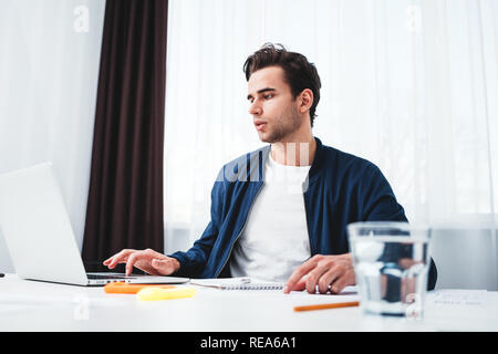 Concentrate l uomo la scrittura di nuove idee in notepad guardando sullo schermo portatile. Giovane collega che siede a tavola e lavoro in ufficio Foto Stock