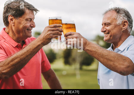 Due alti uomini tostare bicchieri da birra all'esterno. Sorridente maschio maturo amici tifo birre mentre in piedi al di fuori. Foto Stock