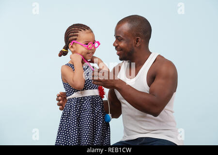 Felice padre e figlia riproduzione medico con stetoscopio giocattolo contro il fondo azzurro. Indossa blue polka dot dress e trecce. Egli è in grassetto, ho Foto Stock