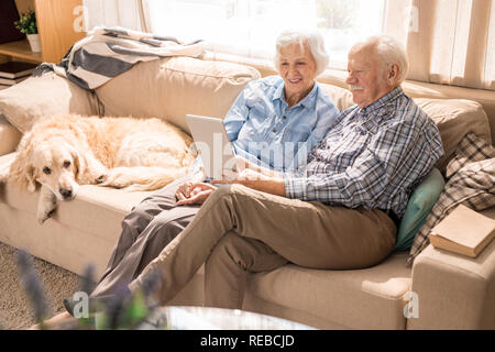 A piena lunghezza ritratto della moderna coppia senior utilizzando computer portatile mentre è seduto con il cane sul lettino a casa Foto Stock