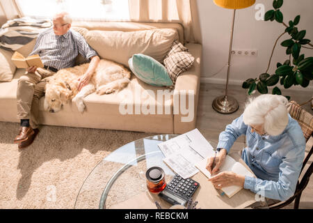 Angolo alto ritratto di donna senior il riempimento di forme e di pagare le tasse con uomo seduto sul divano con il cane, spazio di copia Foto Stock