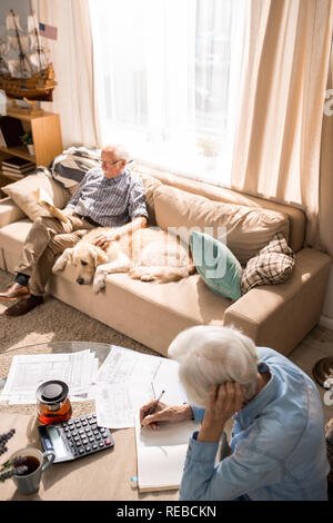 Angolo alto ritratto dell uomo vecchio con il cane seduto sul divano in presenza di luce solare con il senior donna riempire moduli fiscali in primo piano, spazio di copia Foto Stock