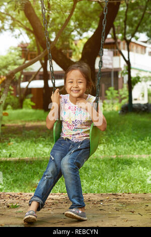 Sorridente ragazza asiatica seduti sulle altalene nella soleggiata giornata splendente sullo sfondo Foto Stock